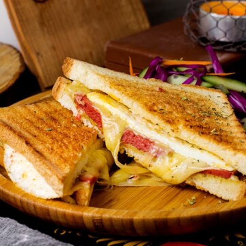 Grilled sandwich with melted cheese and red meat, served with a side salad on a wooden plate.