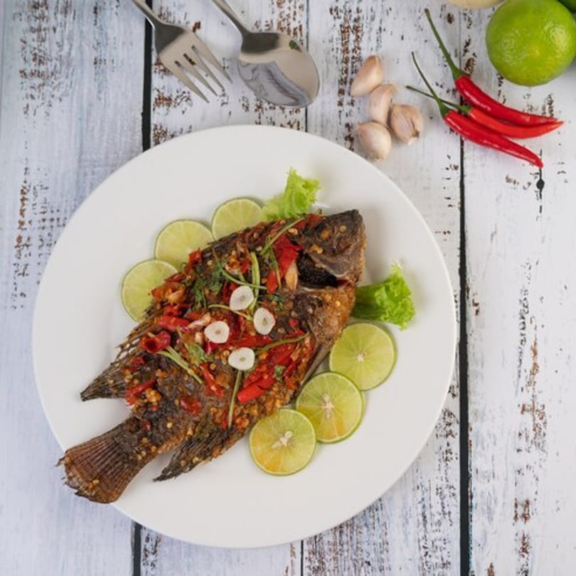 Whole fried fish with chili, garlic, and lime slices on a white plate.